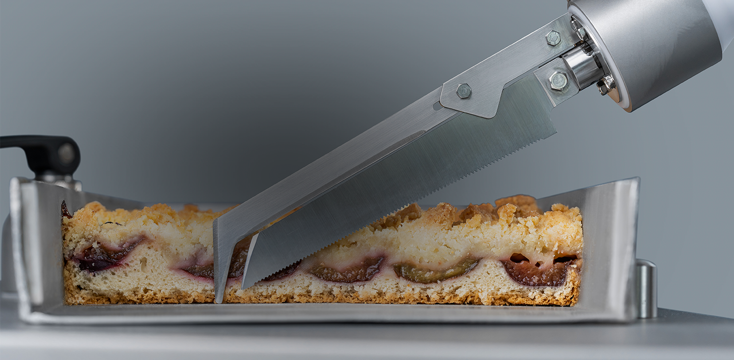 Detailansicht einer pneumatischen Messerklinge, die vor einem Pflaumenkuchen in einem Blech liegt.